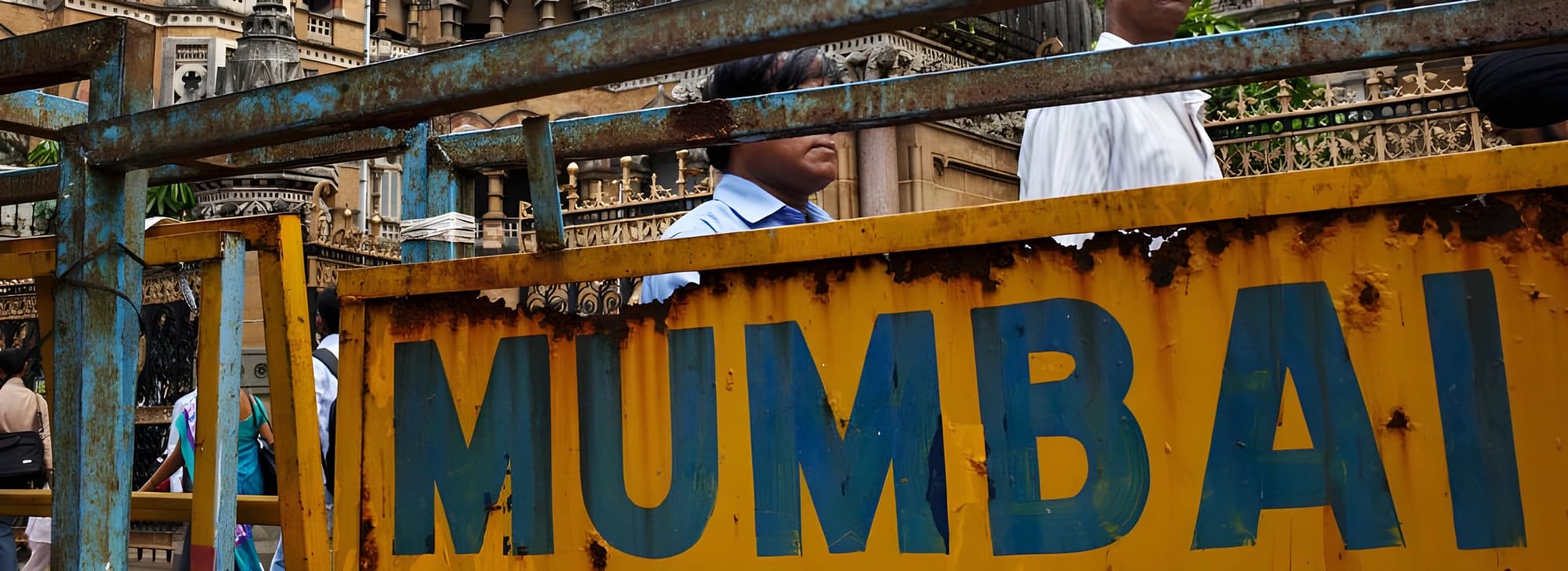 A rusted metal barrier with "Mumbai" partially visible. In the background, people walk past an ornate building.