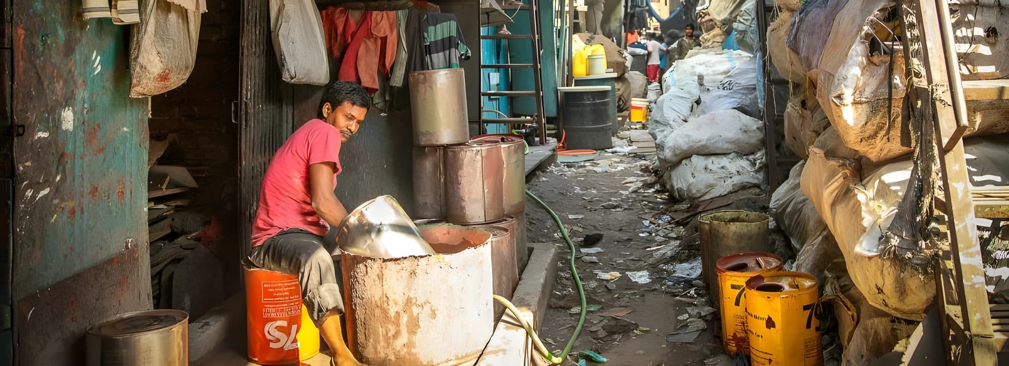 A man stands in a narrow alley surrounded by garbage and pots, highlighting urban neglect and cluttered living conditions.