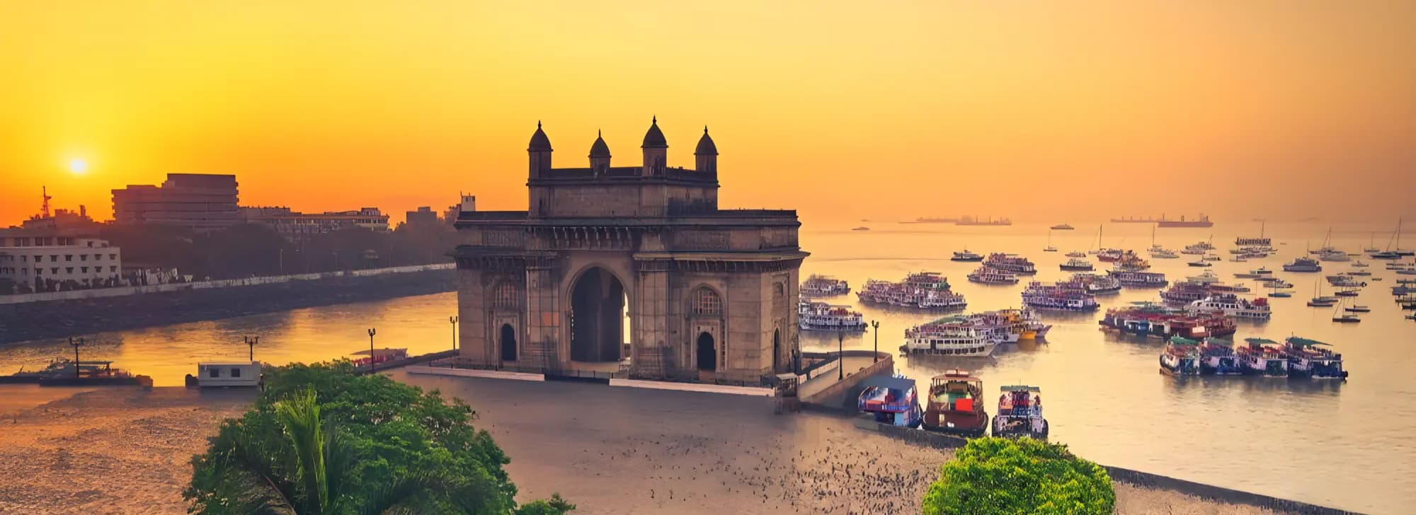 The Gateway of India at sunset, with numerous boats in the water and a cityscape in the background.